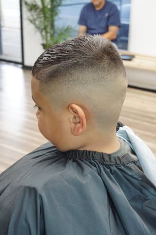 Back view of a young boy with a fade haircut at a barbershop, showing short sides and longer textured top