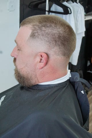 Man with short blonde hair and beard shown in profile, wearing a black cape in a barber shop setting