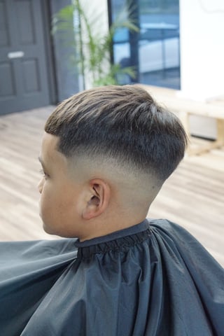 Side profile of a man with a modern fade haircut, short tapered sides and longer textured top, wearing a black cape in an indoor salon setting