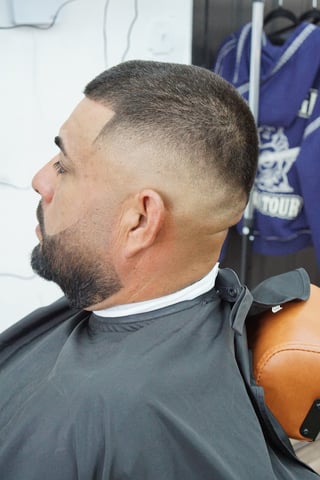 Man with short fade haircut and beard shown in profile, wearing dark cape in barber shop
